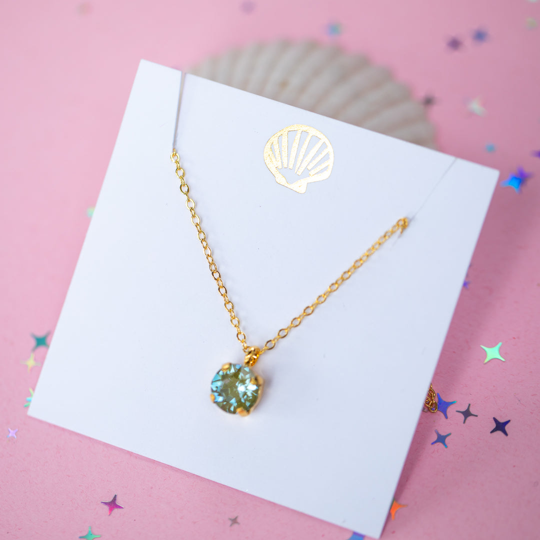 A gold chain necklace with a round Olive green pendant, displayed on a white card with a gold seashell logo, set against a pink background with star confetti.