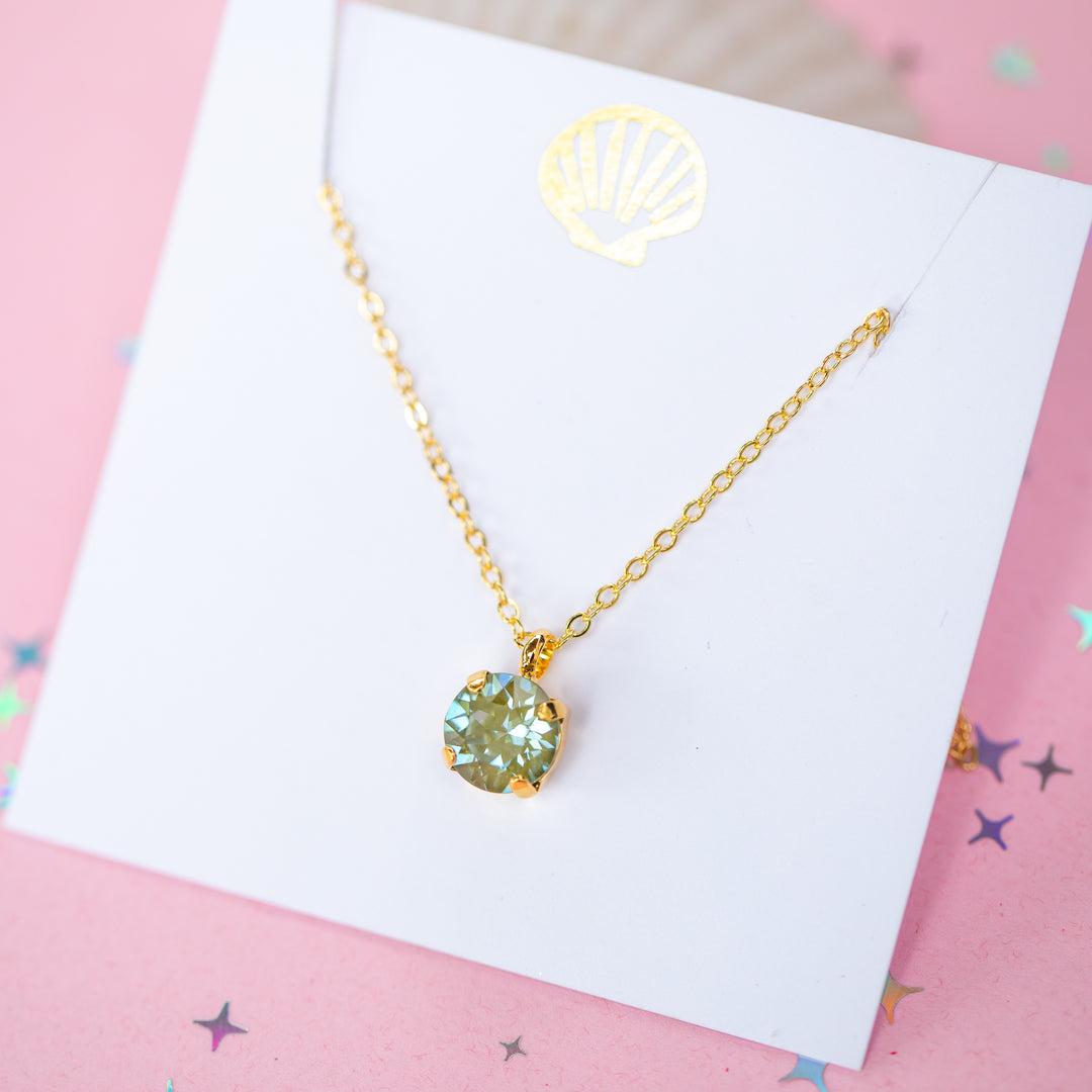 A gold chain necklace with a round Olive green pendant, displayed on a white card with a gold seashell logo, set against a pink background with star confetti.