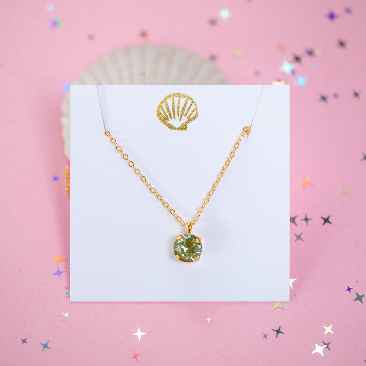 A gold chain necklace with a round Olive green pendant, displayed on a white card with a gold seashell logo, set against a pink background with star confetti.