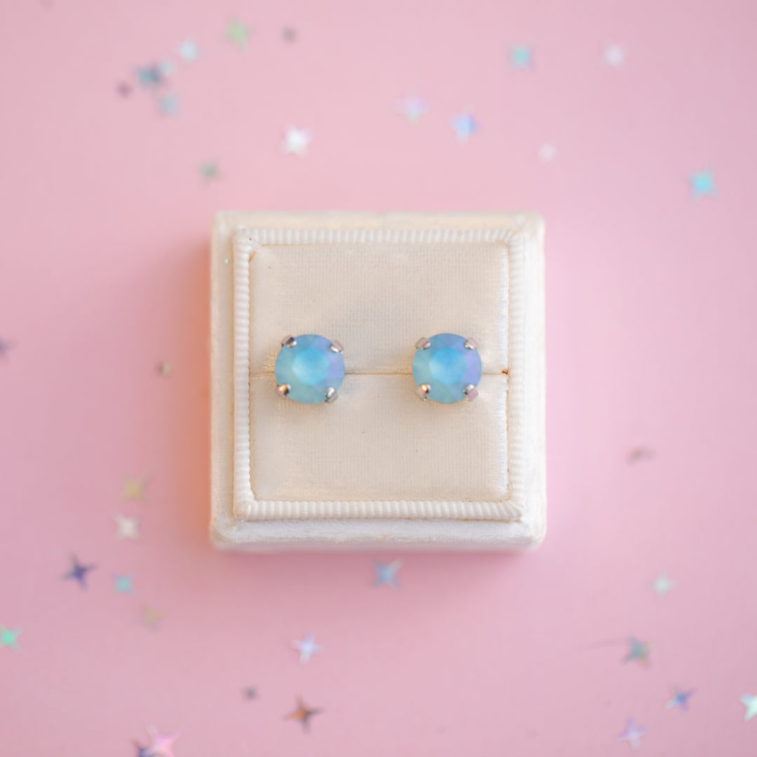 Pair of light blue stud earrings in a white box on a pink background with star patterns.