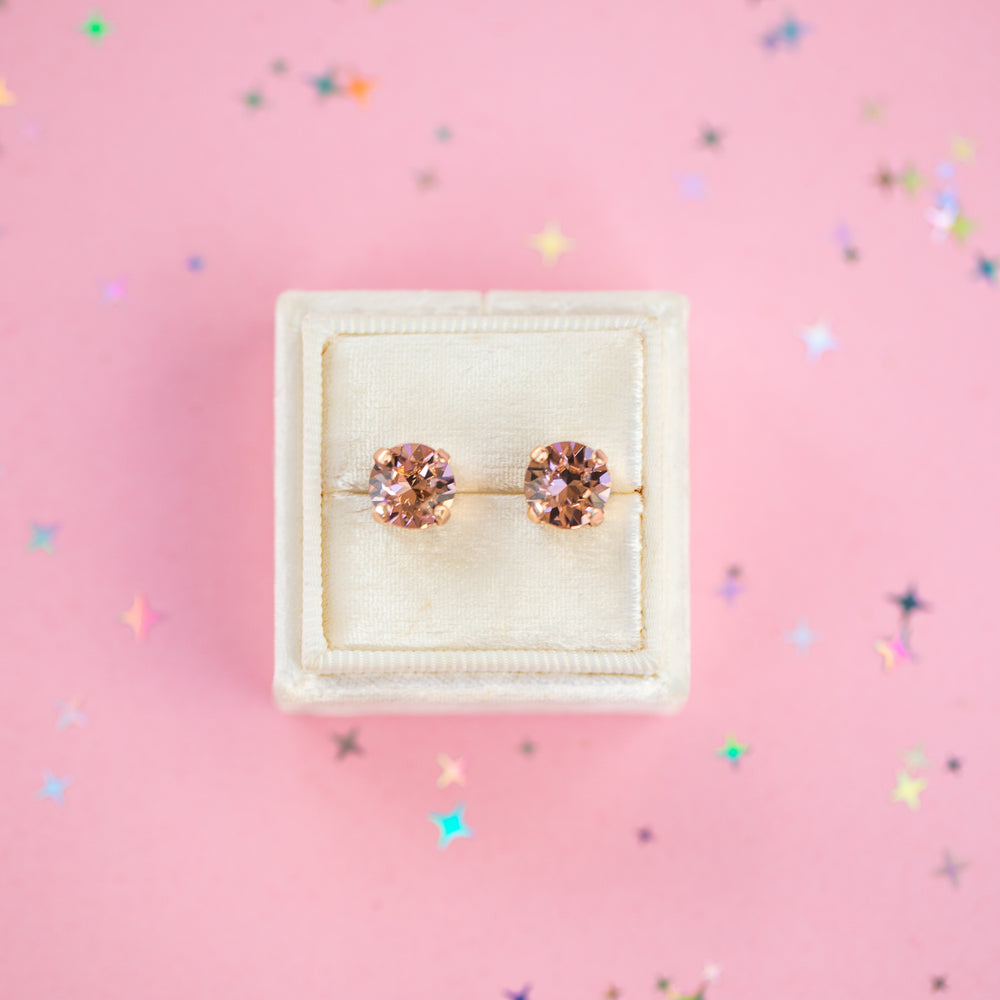 A pair of round-cut rose gold stud earrings with sparkling peach gemstones, presented in a cream-colored jewelry box on a pink background with subtle star confetti.