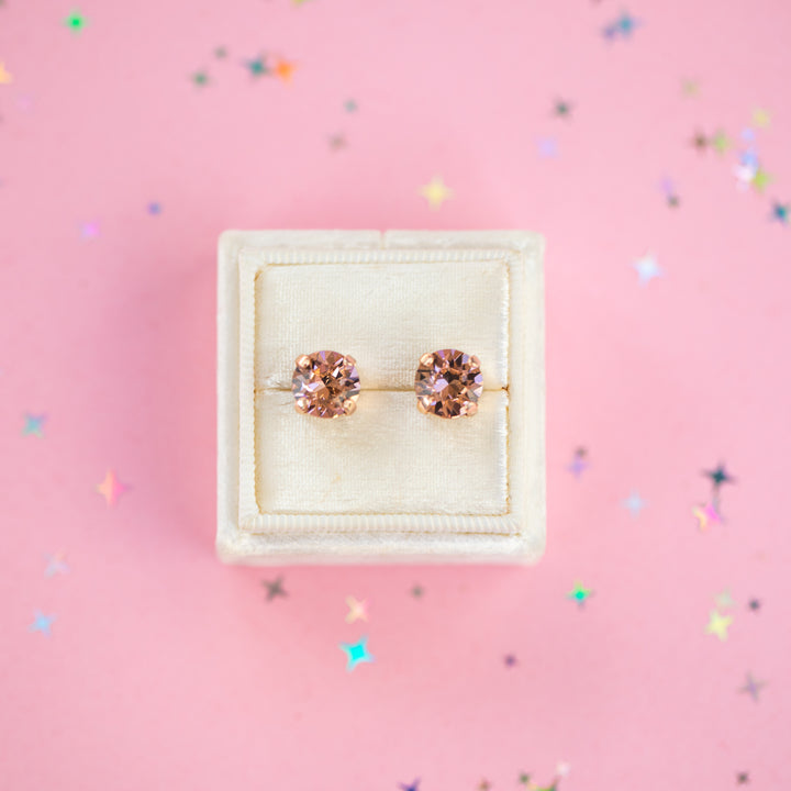 A pair of round-cut rose gold stud earrings with sparkling peach gemstones, presented in a cream-colored jewelry box on a pink background with subtle star confetti.