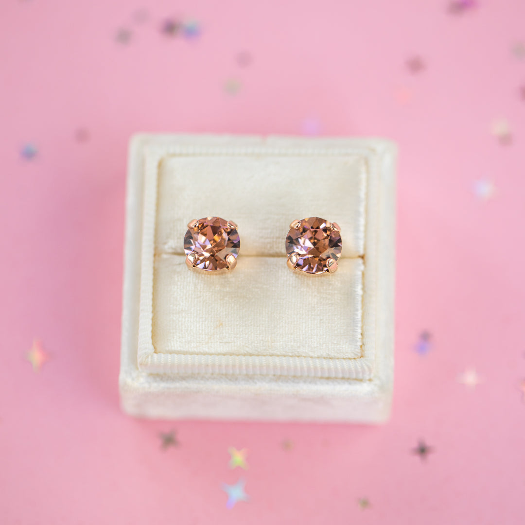 A pair of round-cut rose gold stud earrings with sparkling peach gemstones, presented in a cream-colored jewelry box on a pink background with subtle star confetti.