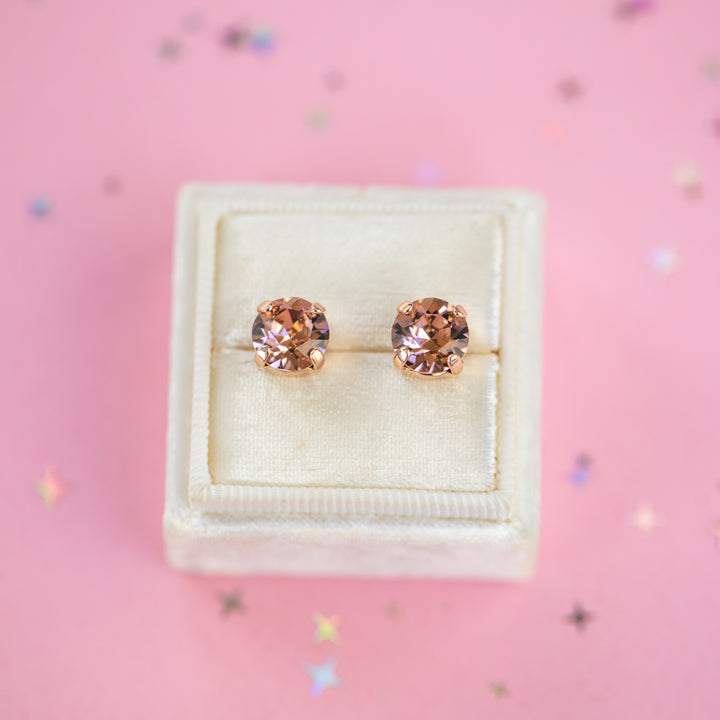A pair of round-cut rose gold stud earrings with sparkling peach gemstones, presented in a cream-colored jewelry box on a pink background with subtle star confetti.