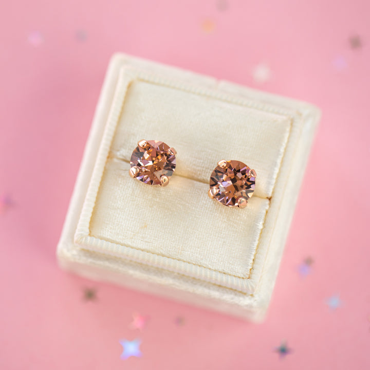 A pair of round-cut rose gold stud earrings with sparkling peach gemstones, presented in a cream-colored jewelry box on a pink background with subtle star confetti.