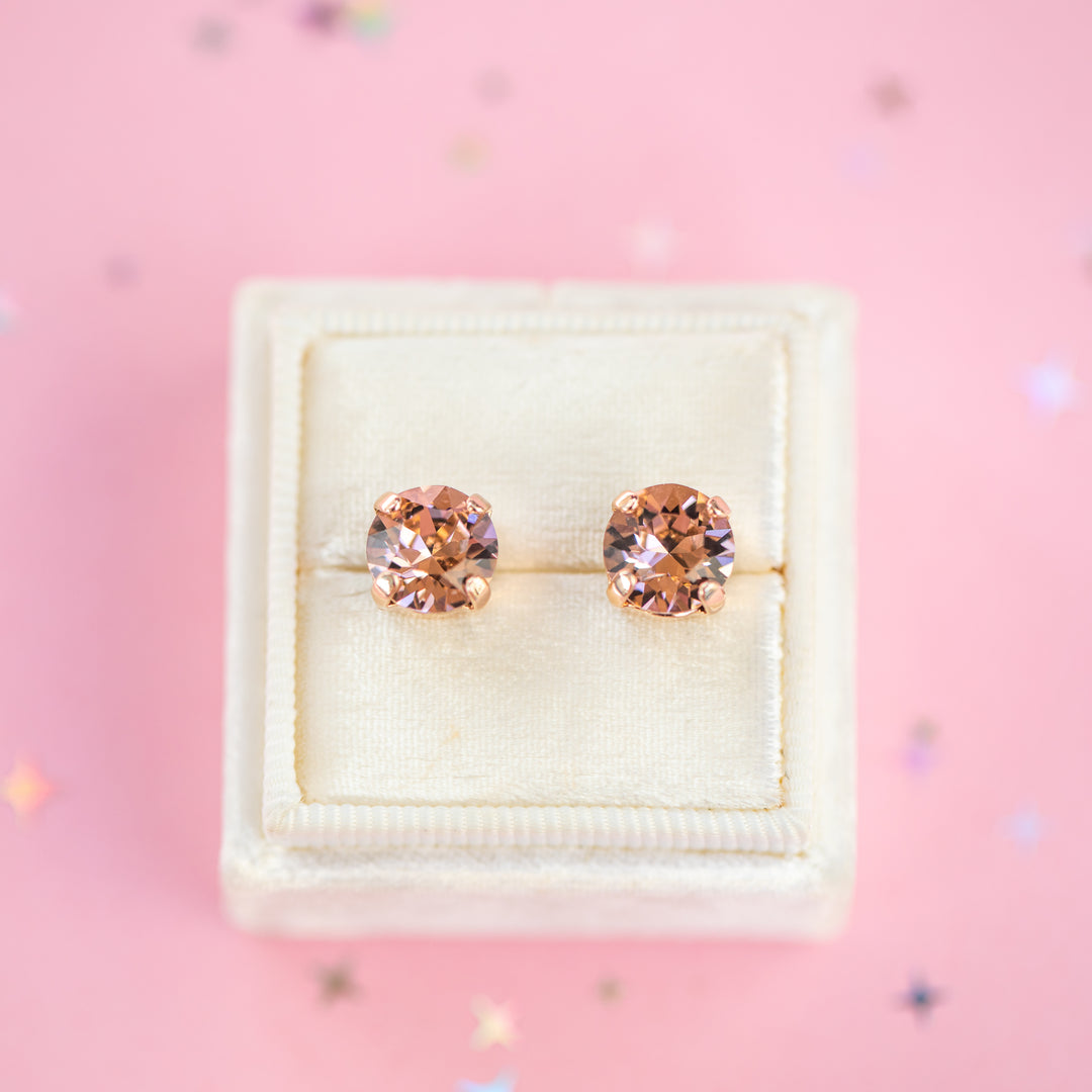 A pair of round-cut rose gold stud earrings with sparkling peach gemstones, presented in a cream-colored jewelry box on a pink background with subtle star confetti.