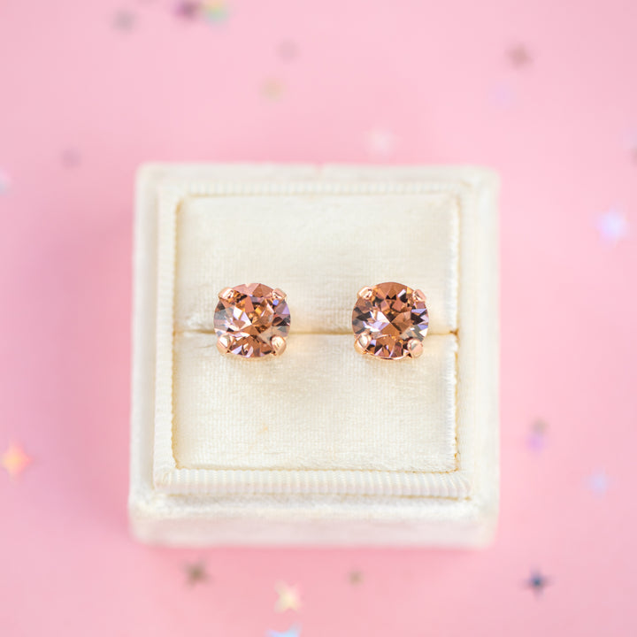 A pair of round-cut rose gold stud earrings with sparkling peach gemstones, presented in a cream-colored jewelry box on a pink background with subtle star confetti.