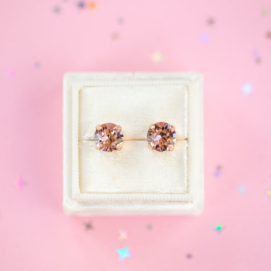 A pair of round-cut rose gold stud earrings with sparkling peach gemstones, presented in a cream-colored jewelry box on a pink background with subtle star confetti.
