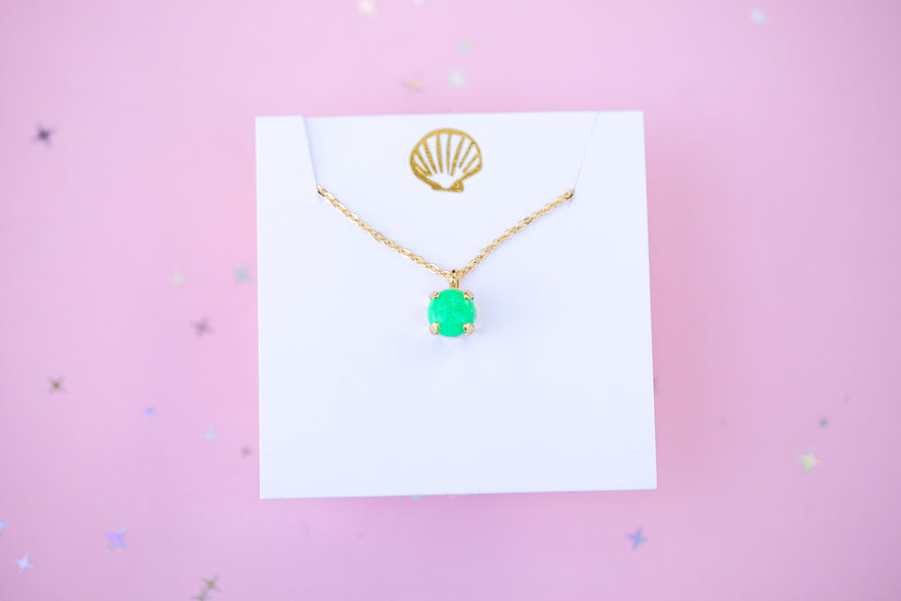 Gold necklace with a green pendant on a white card against a pink background