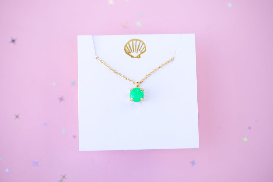 Gold necklace with a green pendant on a white card against a pink background