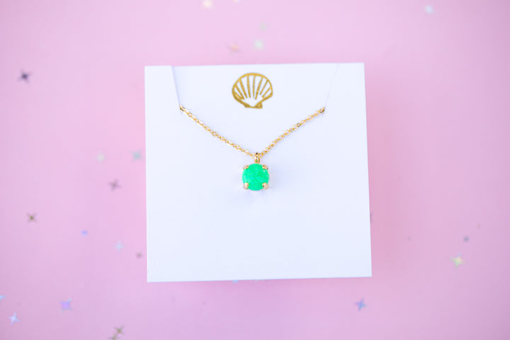Gold necklace with a green pendant on a white card against a pink background