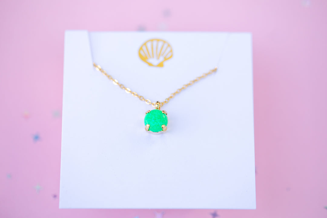 Gold necklace with a green pendant on a white card against a pink background