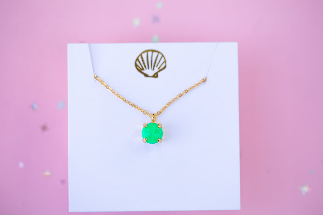 Gold necklace with a green pendant on a white card against a pink background