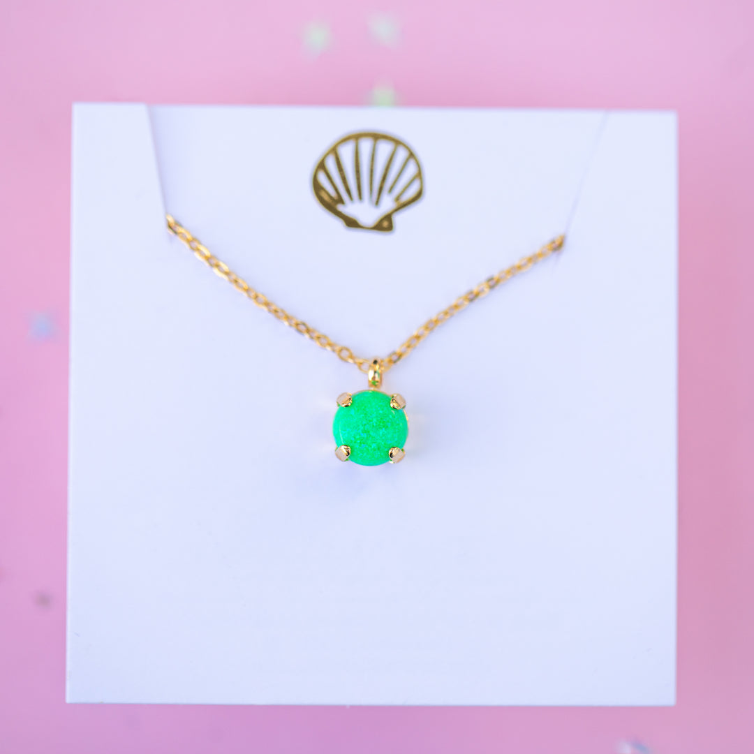 Gold necklace with a green pendant on a white card against a pink background