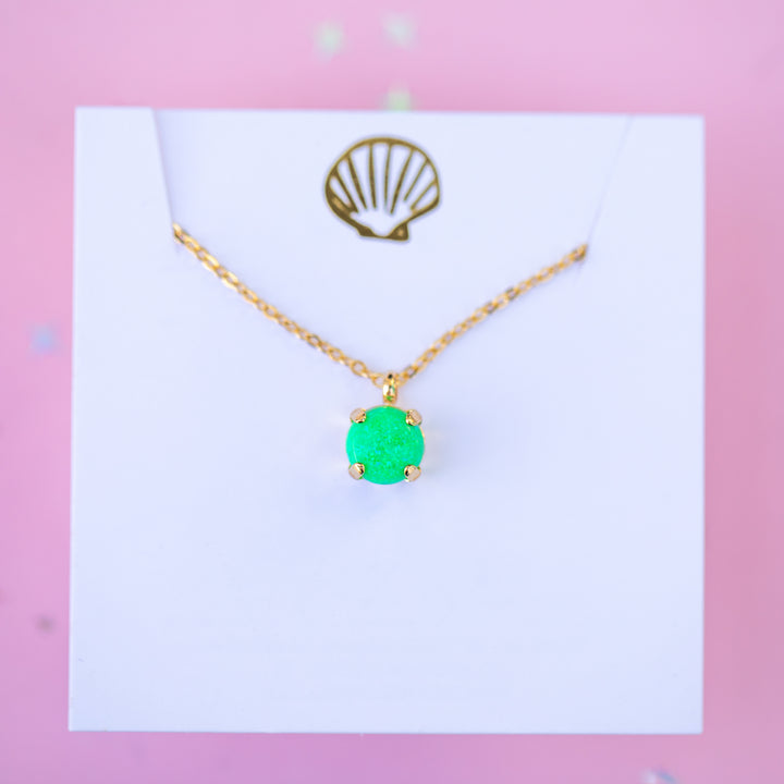 Gold necklace with a green pendant on a white card against a pink background