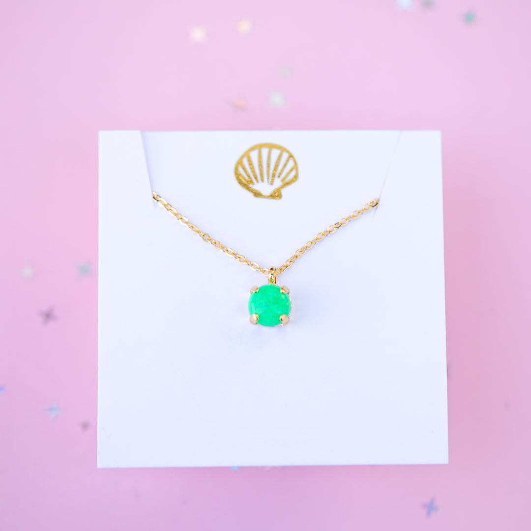 Gold necklace with a green pendant on a white card against a pink background