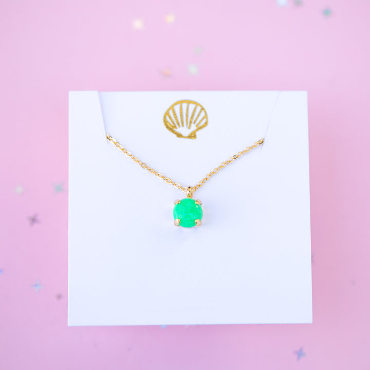 Gold necklace with a green pendant on a white card against a pink background