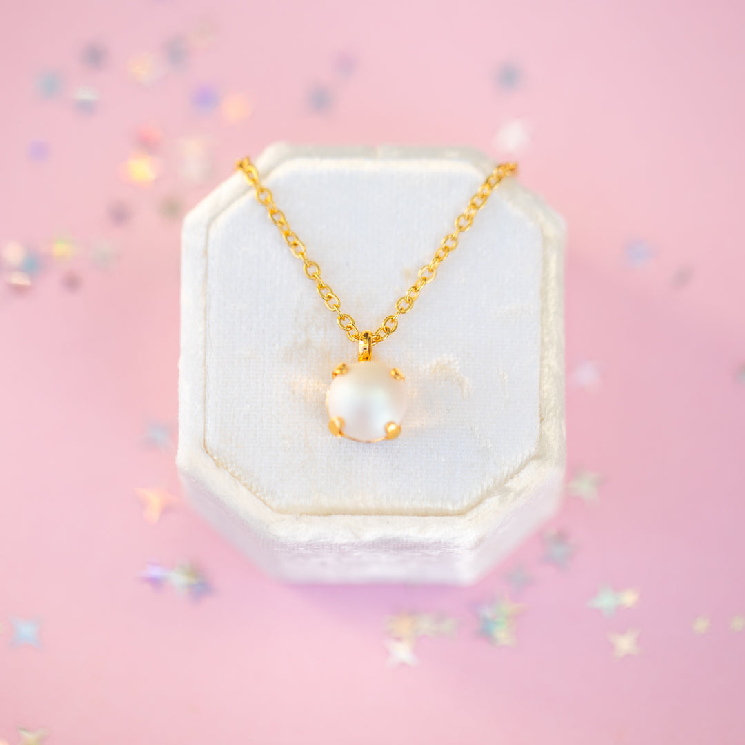 Gold necklace with a pearl pendant on a white hexagonal stand against a pink background