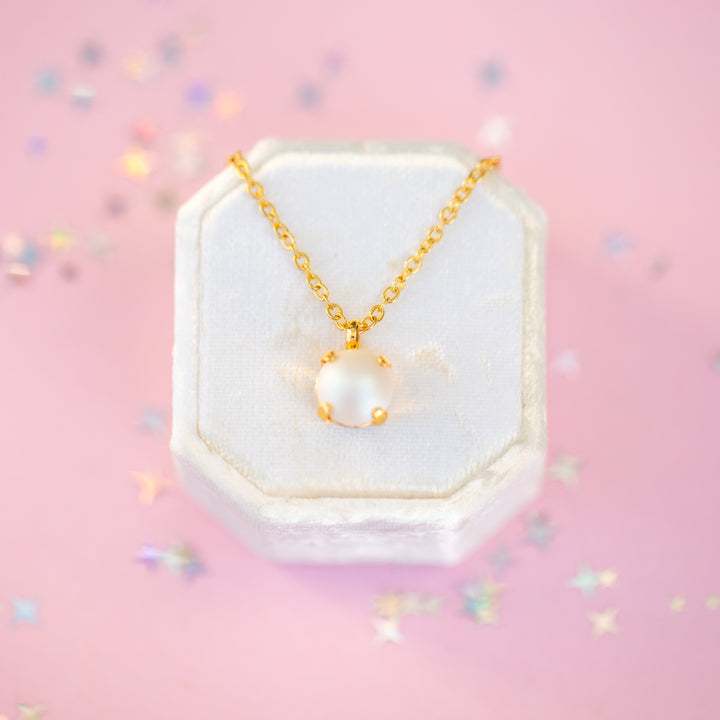 Gold necklace with a pearl pendant on a white hexagonal stand against a pink background