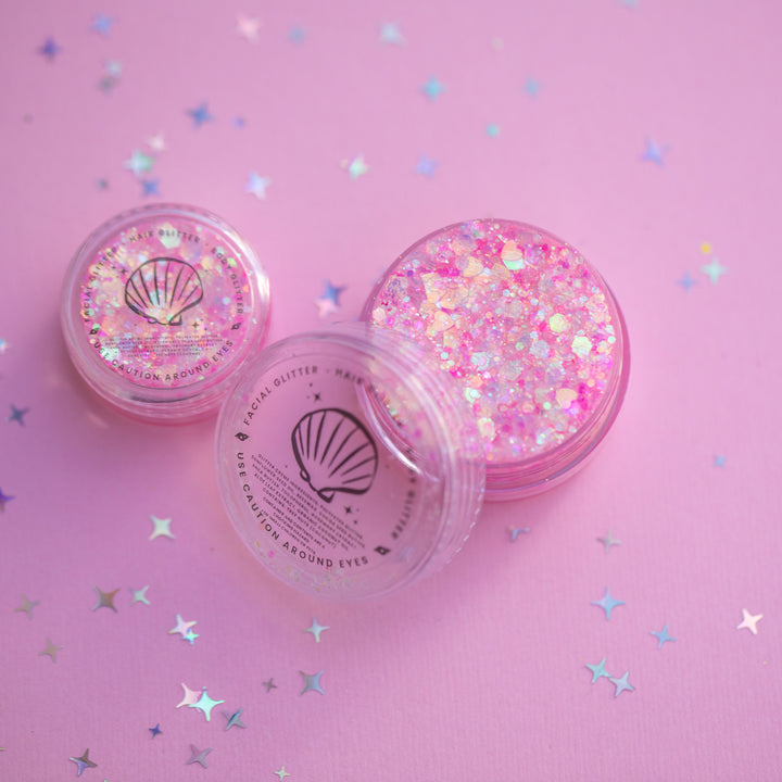 Two containers of glitter with a shell design on a pink background with star-shaped confetti.