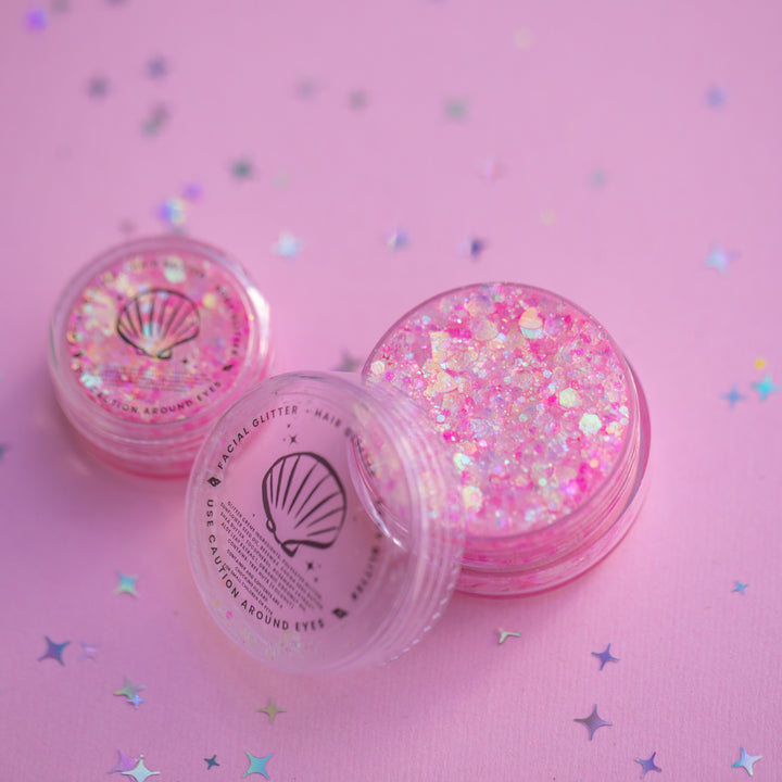 Two containers of pink glitter with a shell design on a pink background with star confetti.