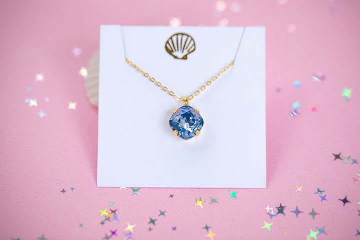 A gold chain necklace with a cushion cut shaped blue faceted gemstone pendant, displayed on a white card with a gold seashell graphic against a pink background.
