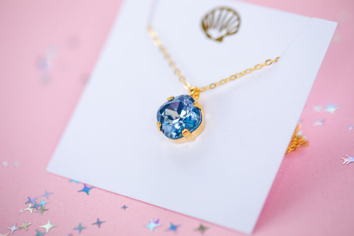 A gold chain necklace with a cushion cut shaped blue faceted gemstone pendant, displayed on a white card with a gold seashell graphic against a pink background.