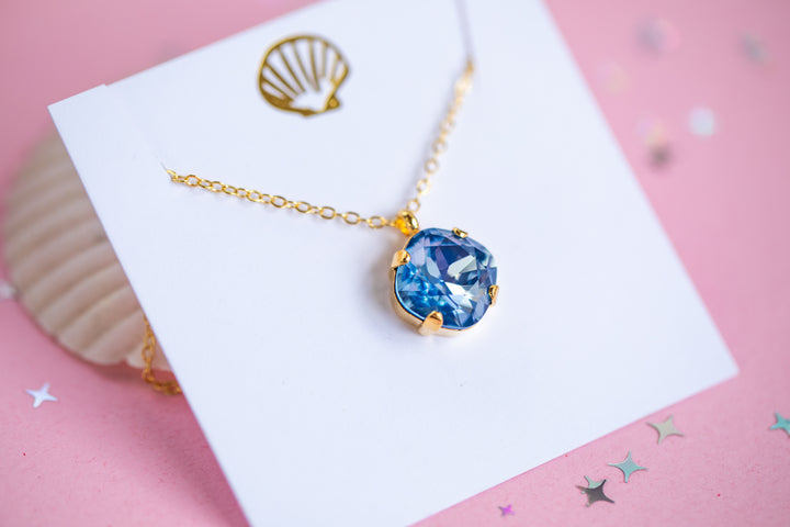 A gold chain necklace with a cushion cut shaped blue faceted gemstone pendant, displayed on a white card with a gold seashell graphic against a pink background.