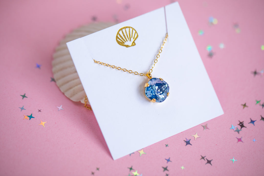 A gold chain necklace with a cushion cut shaped blue faceted gemstone pendant, displayed on a white card with a gold seashell graphic against a pink background.