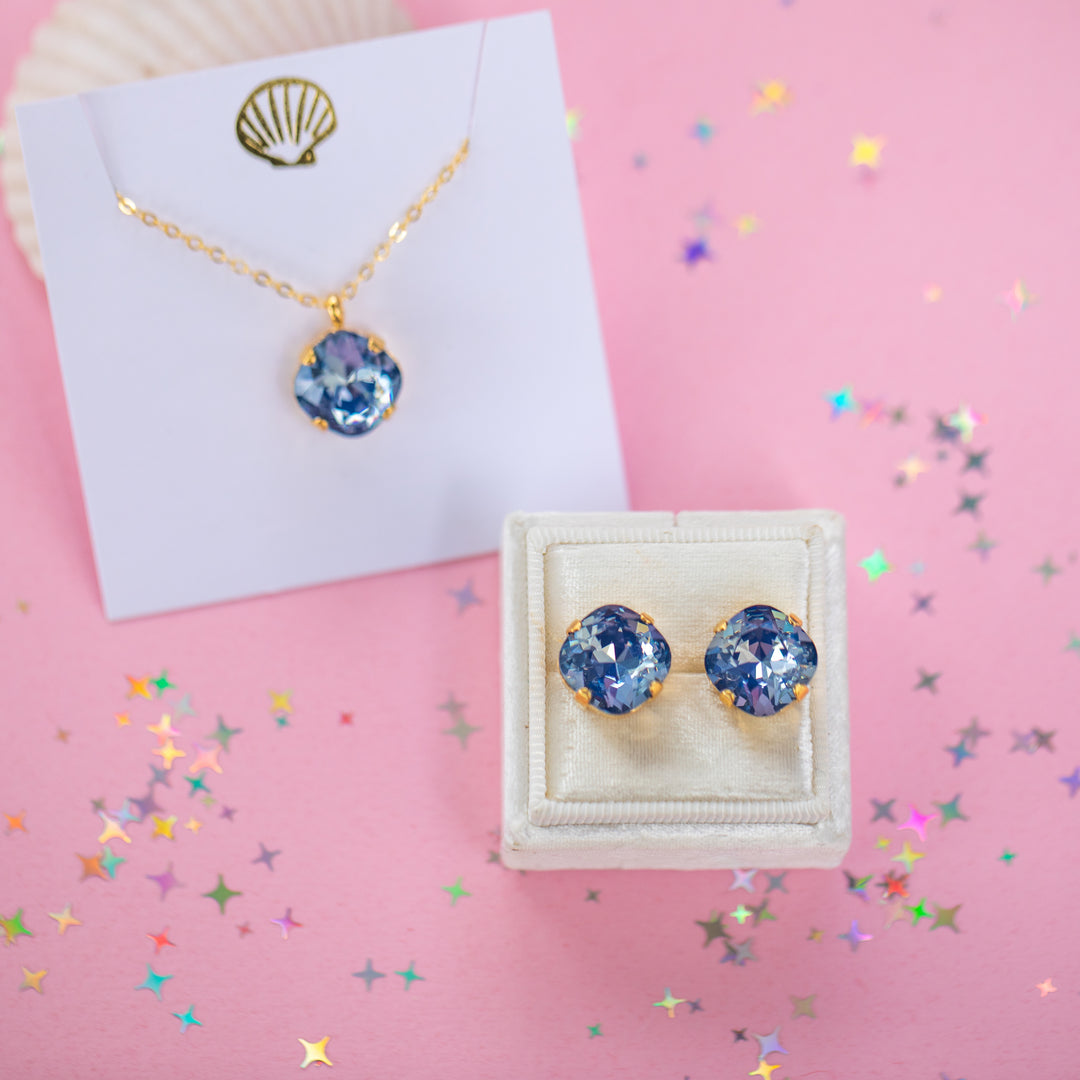 A gold chain necklace and earrings with a cushion cut shaped blue faceted gemstone pendant, displayed on a white card with a gold seashell graphic against a pink background.