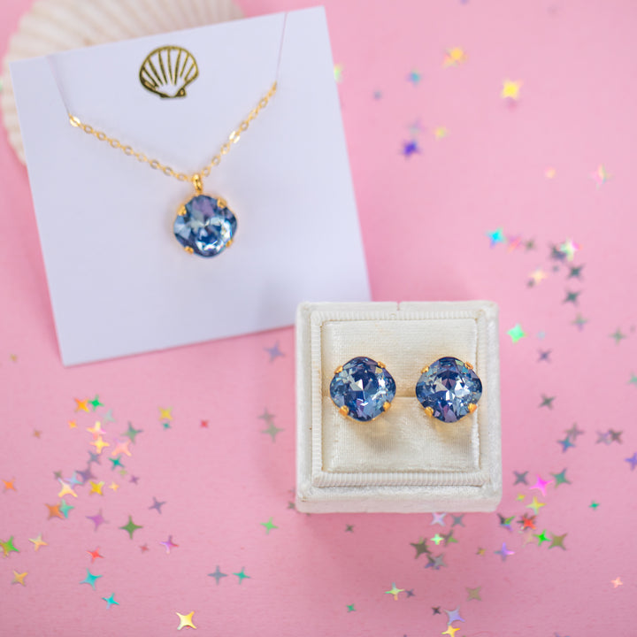 A gold chain necklace and earrings with a cushion cut shaped blue faceted gemstone pendant, displayed on a white card with a gold seashell graphic against a pink background.