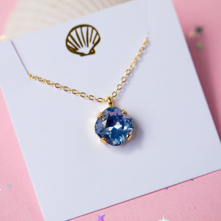 A gold chain necklace with a cushion cut shaped blue faceted gemstone pendant, displayed on a white card with a gold seashell graphic against a pink background.