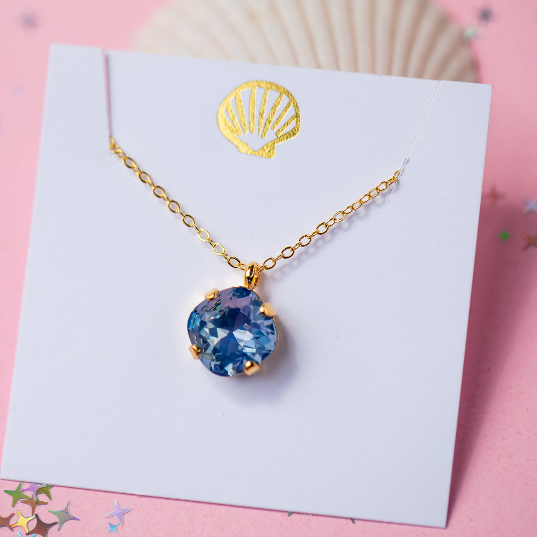 A gold chain necklace with a cushion cut shaped blue faceted gemstone pendant, displayed on a white card with a gold seashell graphic against a pink background.