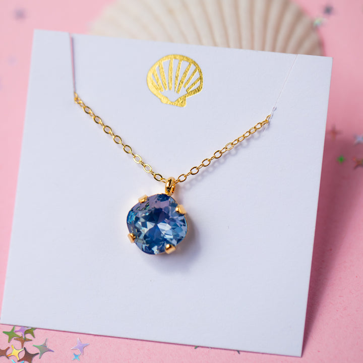 A gold chain necklace with a cushion cut shaped blue faceted gemstone pendant, displayed on a white card with a gold seashell graphic against a pink background.