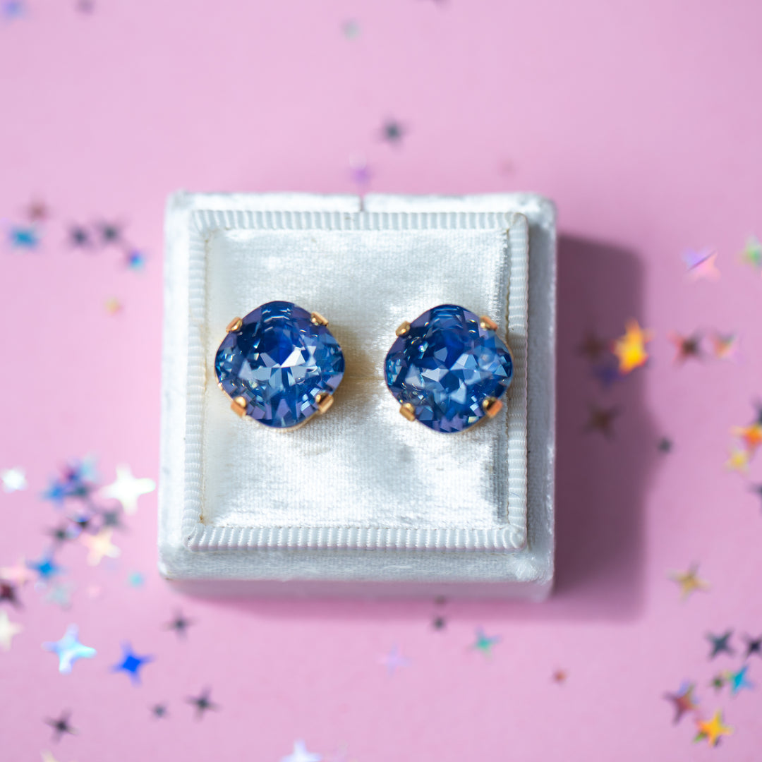 Blue Cushion cut shaped crystals in a stud setting in a white earring box with a pink background