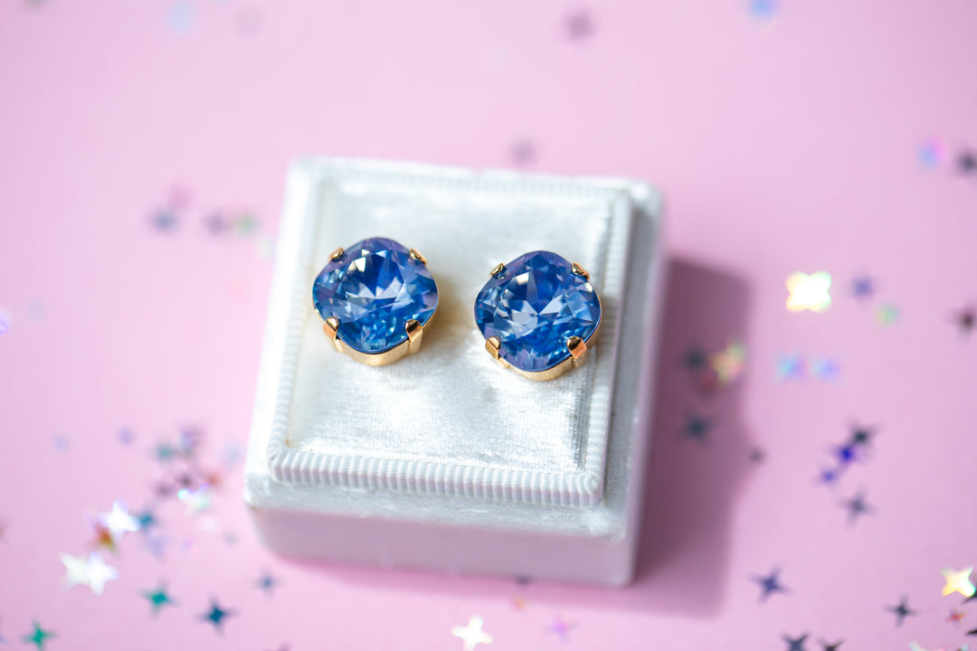 A pair of cushion cut blue gemstone stud earrings in gold settings, displayed on a small white jewelry box against a pink background with scattered star-shaped confetti.