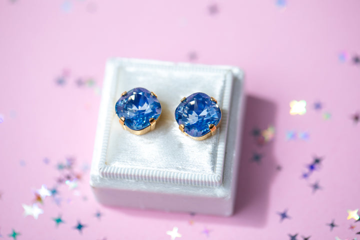 A pair of cushion cut blue gemstone stud earrings in gold settings, displayed on a small white jewelry box against a pink background with scattered star-shaped confetti.