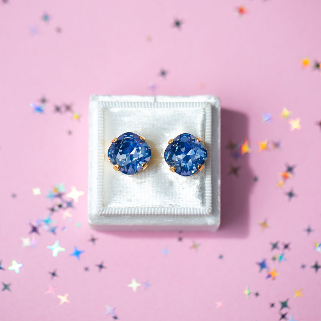 A pair of cushion cut blue gemstone stud earrings in gold settings, displayed on a small white jewelry box against a pink background with scattered star-shaped confetti.