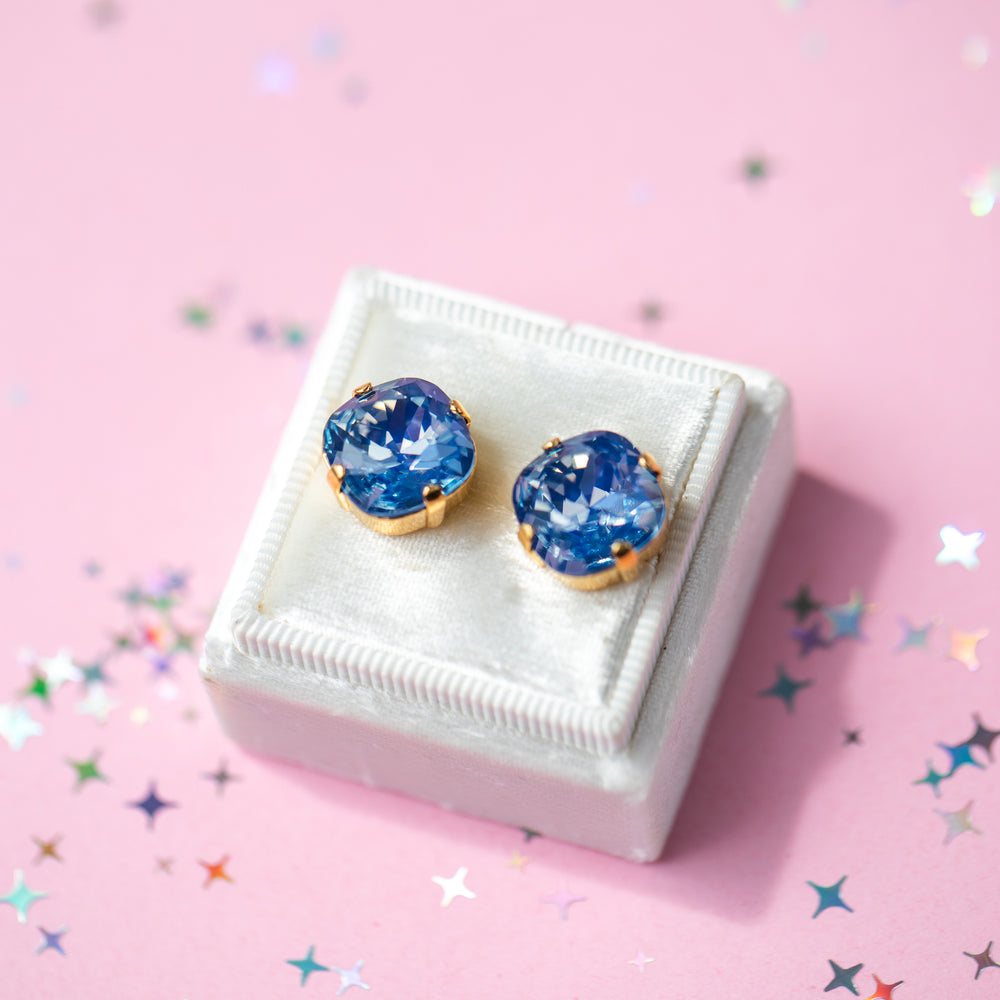 A pair of cushion cut blue gemstone stud earrings in gold settings, displayed on a small white jewelry box against a pink background with scattered star-shaped confetti.