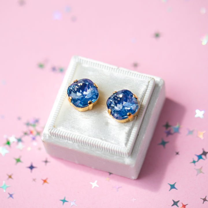 A pair of cushion cut blue gemstone stud earrings in gold settings, displayed on a small white jewelry box against a pink background with scattered star-shaped confetti.