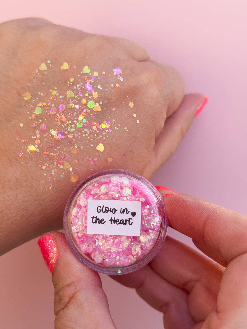 Hand holding a container of pink glitter with 'Glow in the Heart' label against a pink background