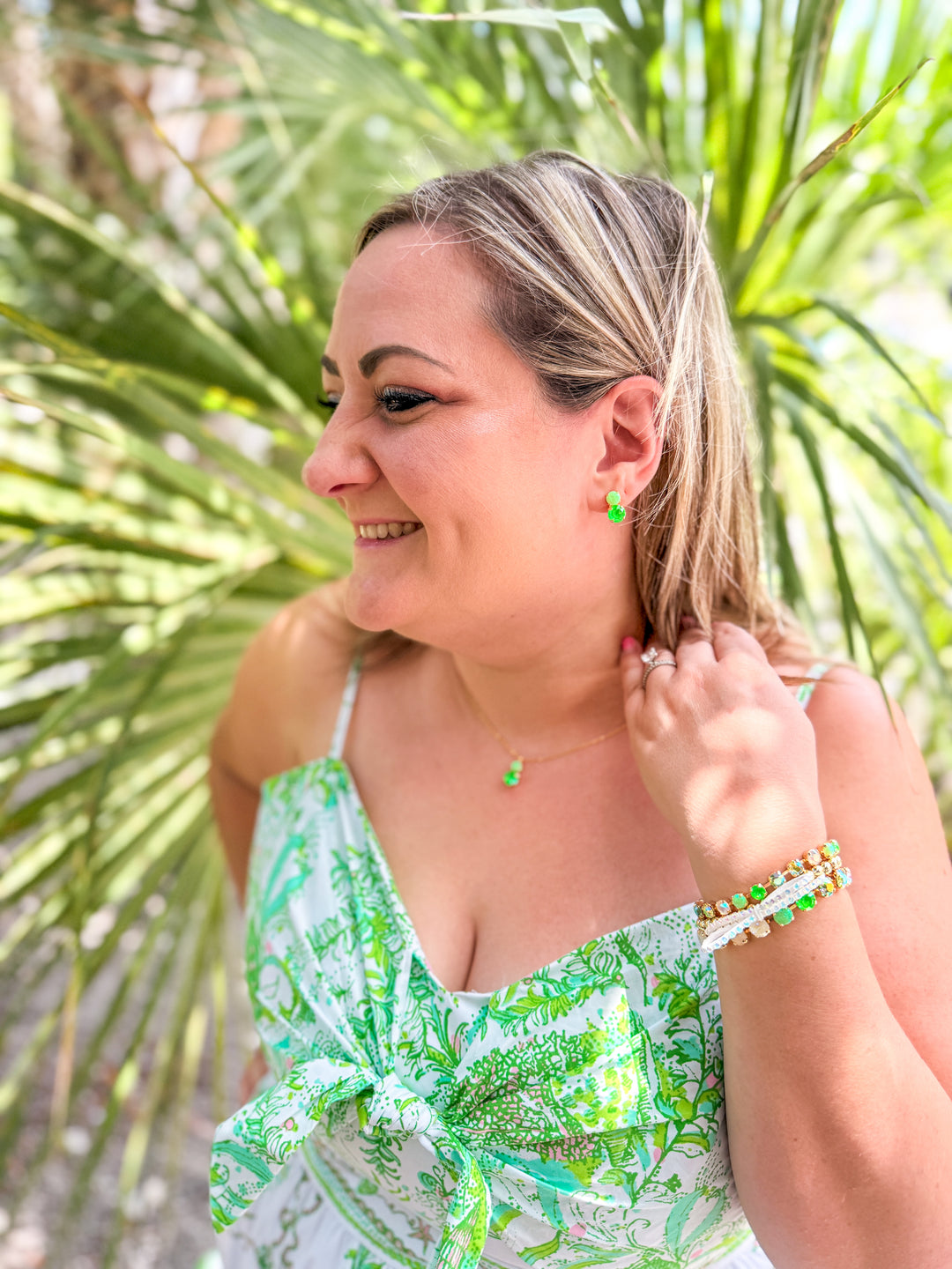 Woman wearing a green and white patterned dress with jewelry, standing in front of palm leaves.