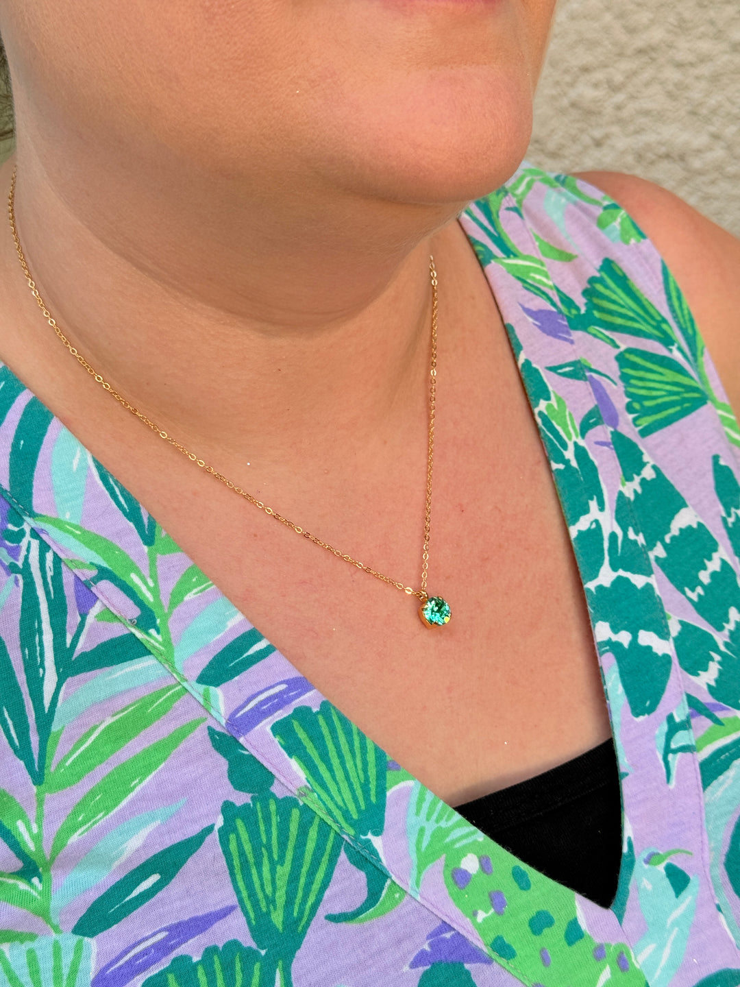 Close-up of a person wearing a gold necklace with a green gemstone, against a tropical patterned background.