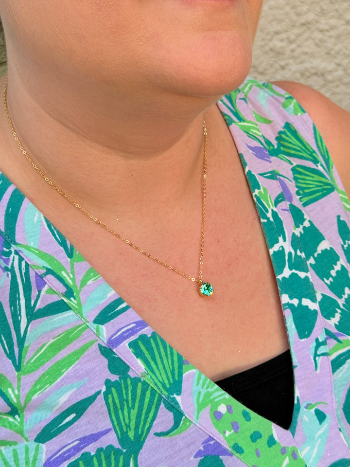 Close-up of a person wearing a gold necklace with a green gemstone, against a tropical patterned background.