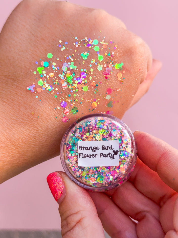Multicolored glitter in a small round container held by a hand with pink nail polish on a pink background.