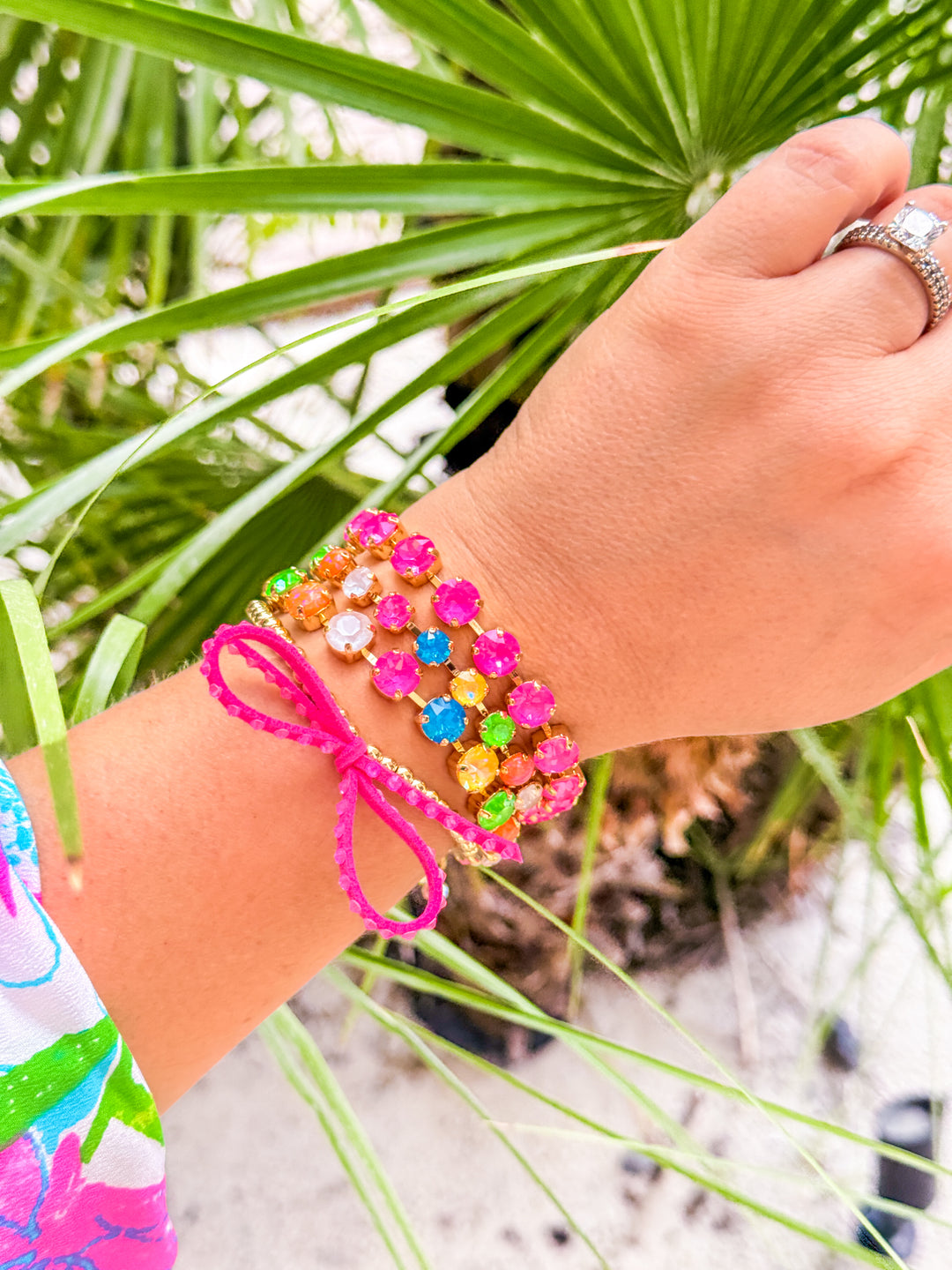 A close-up of a person's wrist wearing a colorful beaded bracelet with a bright pink braided cord and bow, set against a background of green palm leaves and white sand.