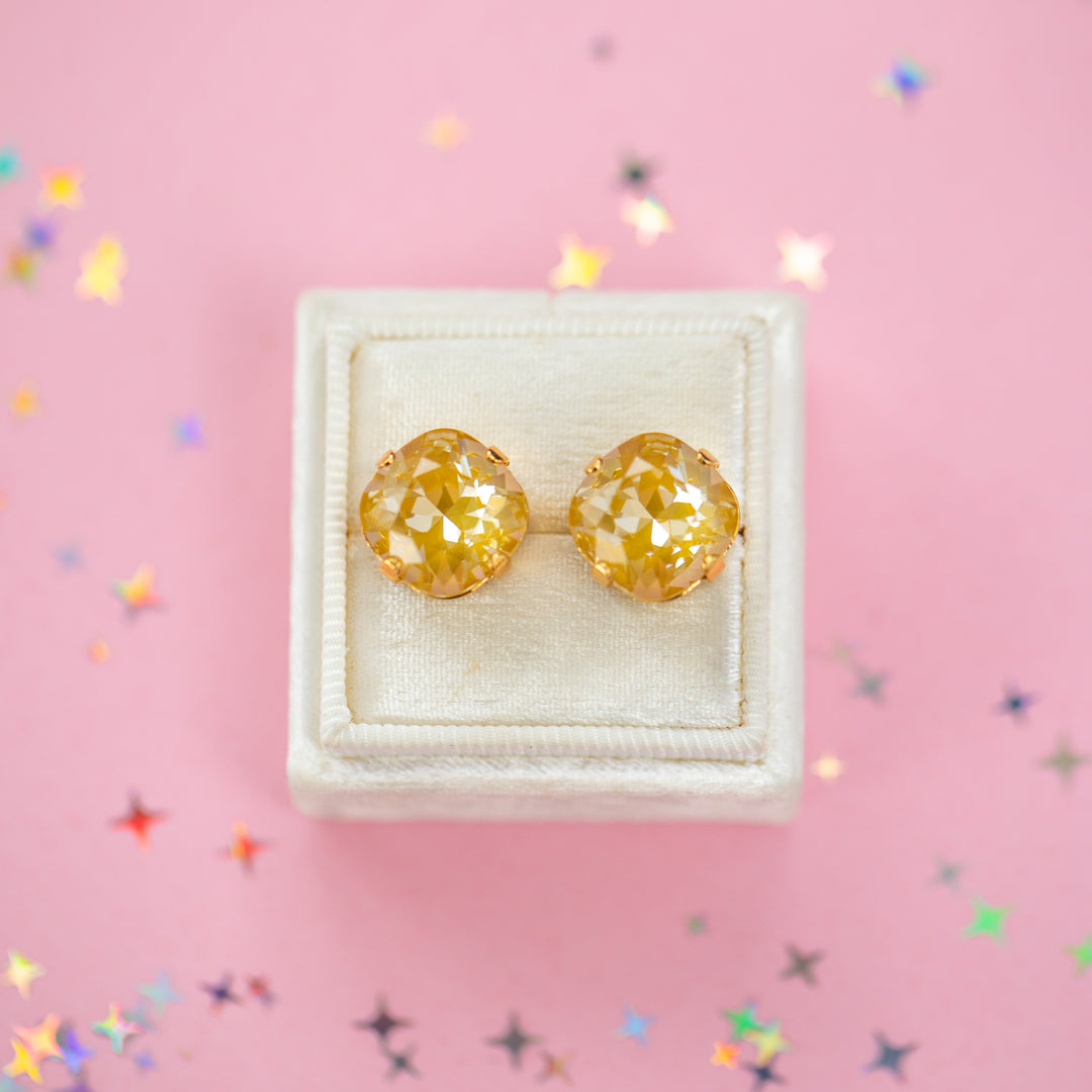 A pair of mustard yellow faceted cushion cut stud earrings displayed on a cream-colored jewelry box against a pink background with scattered star-shaped confetti.