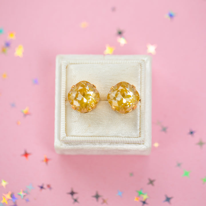 A pair of mustard yellow faceted cushion cut stud earrings displayed on a cream-colored jewelry box against a pink background with scattered star-shaped confetti.
