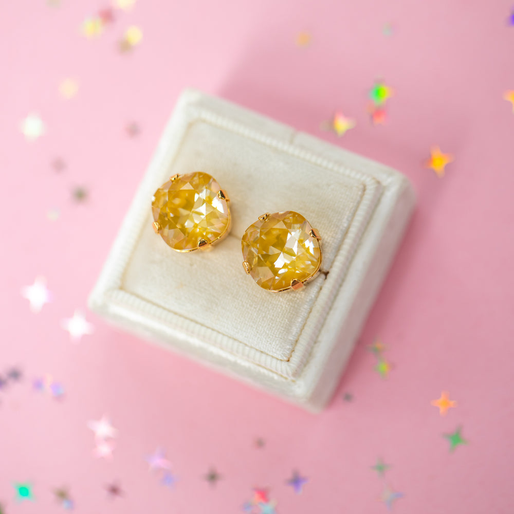 A pair of mustard yellow faceted cushion cut stud earrings displayed on a cream-colored jewelry box against a pink background with scattered star-shaped confetti.