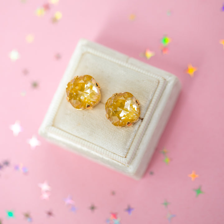 A pair of mustard yellow faceted cushion cut stud earrings displayed on a cream-colored jewelry box against a pink background with scattered star-shaped confetti.